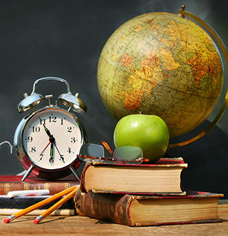 An apple, clock, globe, glasses, stacked books and pencils sit on a table in front of a blackboard